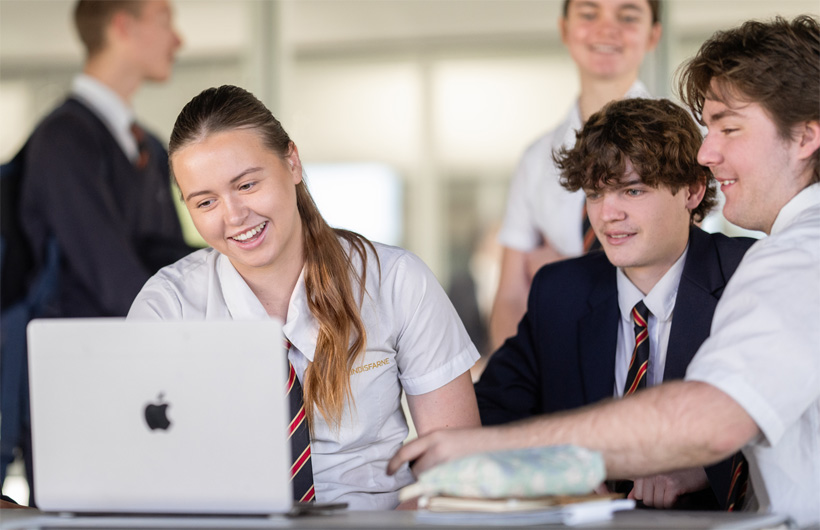 Un groupe d’élèves en uniforme, le sourire aux lèvres, est rassemblé autour d’un MacBook pour collaborer.