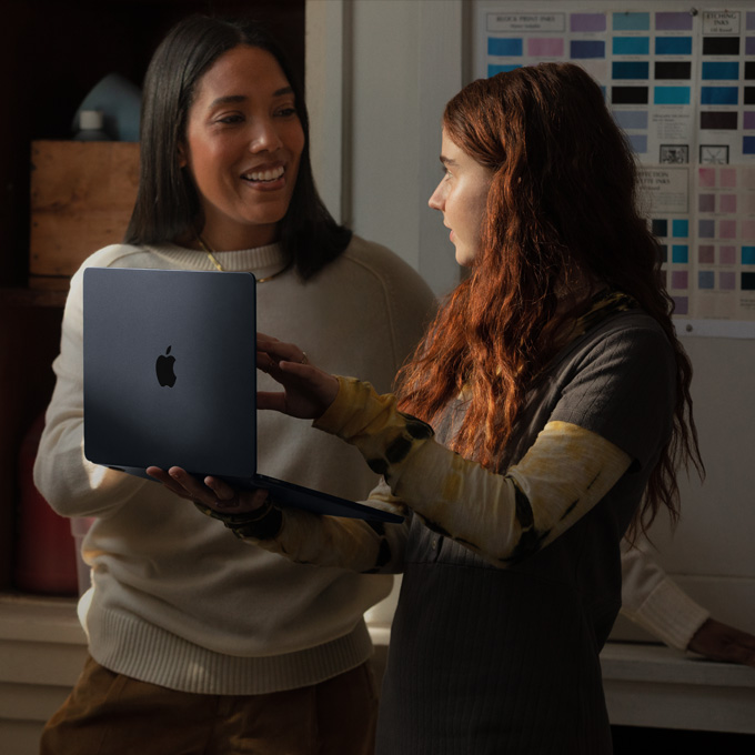 Una profesora y una estudiante conversan mientras sostienen un notebook MacBook