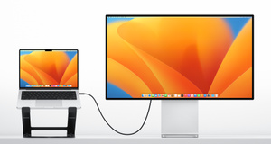 The Curve Flex Stand, in fully raised position, with open MacBook on top of it, cable-connected to a mirrored large-screen monitor.
