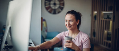A woman sits at a desk, holding a mug in one hand and using a large computer monitor with the other