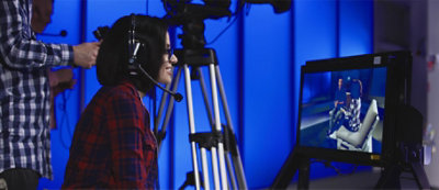 Person wearing a headset sits in front of a monitor with a video camera on a tripod directed at them in a blue-lit room.
