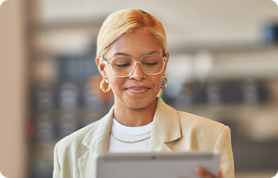 A person with short blonde hair and glasses is looking at a tablet. They are wearing a beige blazer and loop earrings.