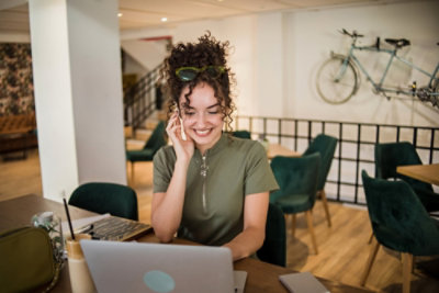 Person using a laptop in a café, on a phone call, with a drink on the table and a bicycle on the wall behind.