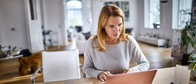 A woman with long hair sits at a table in a bright, modern room, focusing on a laptop in front of her.