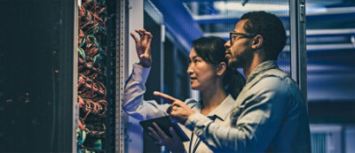 Two individuals are examining a server rack in a data center, pointing out details and discussing while one holds a tablet.