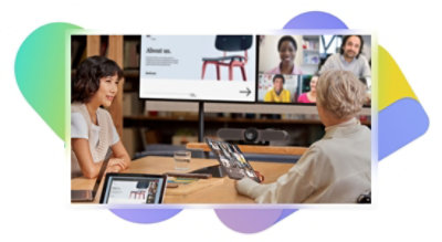Two people in a meeting with large screens displaying a Teams video call and a presentation, one is speaking, and the other is holding a tablet.