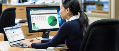 A woman sits at a desk in an office, working on a laptop while a computer monitor in front of her displays a spreadsheet with a green pie chart.