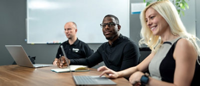 Three people are seated at a conference table, each with laptops; one person in a black shirt is holding a pen and looking towards the camera
