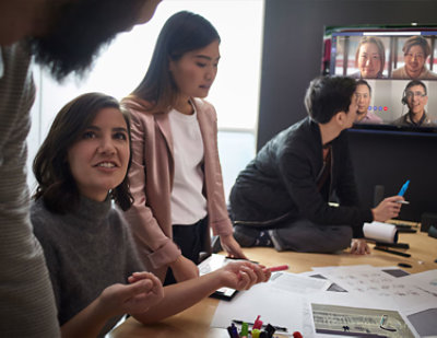 Four people collaborating in a meeting room with documents on table; three engage while one joins via video call 