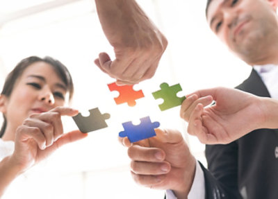 Four people hold colored puzzle pieces (black, red, green, and blue) and attempt to fit them together