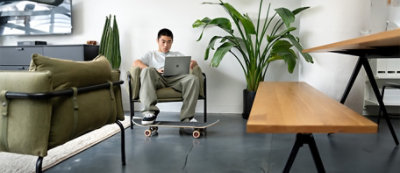 A person sits on a chair in a modern living room, using a laptop placed on their lap. A skateboard rests under their feet, and plants are visible in the background.