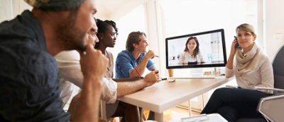 Four people are sitting at a table, attentively looking at a computer screen displaying a video call with a person.