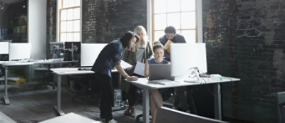 A group of four people are gathered around a laptop in a room with large windows and a textured wall.